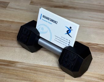 May include: A black dumbbell-shaped business card holder with a silver handle. The ends of the dumbbell are marked with the number 25. A stack of business cards is held in the centre, with the name "RICHARD SANCHEZ, Personal Trainer" visible.