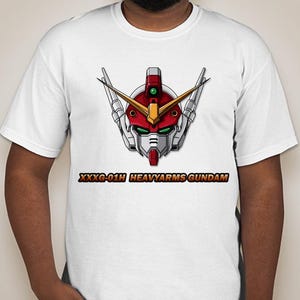 Pu&ograve; includere: Maglietta bianca con un'immagine di una testa di robot rosso, bianco e verde e il testo "XXXG-01H HEAVYARMS GUNDAM".