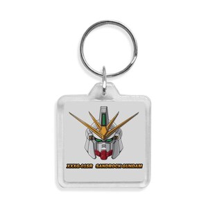 Puede incluir: Un llavero cuadrado con una ilustraci&oacute;n de dibujos animados de una cabeza de robot Gundam. La cabeza del robot es principalmente blanca, con detalles rojos, verdes y dorados. El texto "XXXG-01SR SANDROCK GUNDAM" est&aacute; impreso debajo.