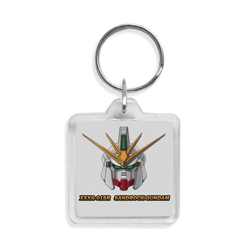 Puede incluir: Un llavero cuadrado con una ilustraci&oacute;n de dibujos animados de una cabeza de robot Gundam. La cabeza del robot es principalmente blanca, con detalles rojos, verdes y dorados. El texto "XXXG-01SR SANDROCK GUNDAM" est&aacute; impreso debajo.