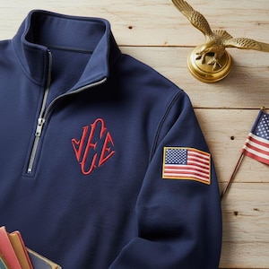 Könnte beinhalten: Marineblaues Sweatshirt mit Viertelreißverschluss, rotem, gesticktem Monogramm und einem amerikanischen Flaggenaufnäher auf dem Ärmel. Das Sweatshirt hat einen silbernen Reißverschluss und einen Stehkragen. Eine kleine amerikanische Flagge und eine goldene Adlerstatue sind ebenfalls zu sehen.