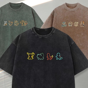 Może przedstawiać: Trzy koszulki z okrągłym dekoltem w odcieniach zieleni, brązu i czerni. Każda koszulka ma kolorowy haftowany wzór postaci z kreskówek. Koszulki mają wygląd vintage.