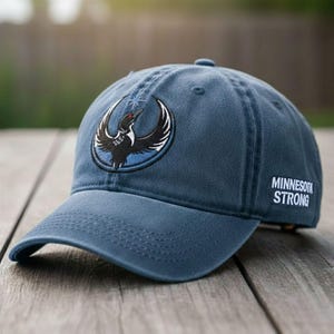 May include: A blue baseball cap with a curved brim. The cap features an embroidered logo of a bird with outstretched wings inside a circle. The text "MINNESOTA STRONG" is embroidered on the side.