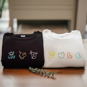 May include: Two sweatshirts, one black and one white, are displayed on a wooden surface. The black sweatshirt features three embroidered Pokémon characters in neon colors. The white sweatshirt has four embroidered Pokémon characters in yellow, green, orange, and blue.