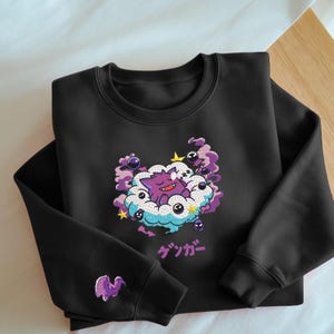 May include: Black sweatshirt featuring a purple cartoon character sleeping on a cloud with purple and blue accents. The Japanese word "ゲンガー" is printed below the graphic. A small purple graphic is on the left sleeve.