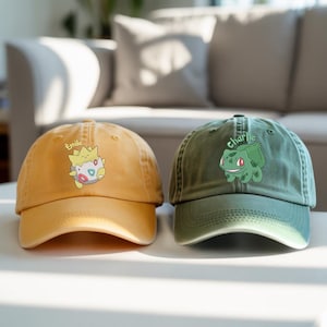 Puede incluir: Dos gorras de béisbol. Una es de color amarillo mostaza con un personaje de dibujos animados y el nombre "Emile". La otra es verde oliva con un personaje de dibujos animados y el nombre "Charlie". Ambas gorras tienen una visera curva.