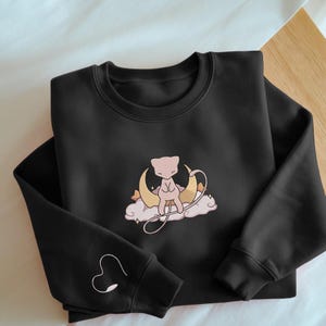 Könnte beinhalten: Schwarzes Sweatshirt mit einem gestickten Design einer rosa Kreatur auf einem Mond und einer Wolke. Auf dem linken Ärmel ist ein Herz gestickt. Das Sweatshirt ist aus weichem Material.