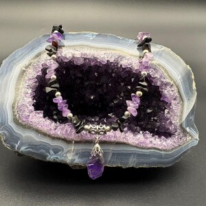 May include: A purple amethyst gemstone necklace with black onyx beads and a silver pendant. The necklace is displayed on a purple amethyst geode.