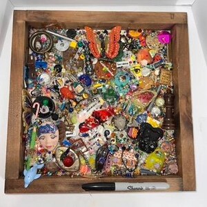 May include: A square wooden shadow box frame filled with a dense collage of colourful trinkets, including a prominent orange butterfly, a doll face, a blue dolphin, a chess piece, beads, and various small charms, with a black Sharpie marker at the base.