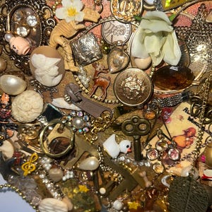 Puede incluir: Un ensamblaje en forma de corazón de joyas y baratijas vintage en tonos dorados, bronce y crema. Incluye botones, dijes, perlas, un dinosaurio en miniatura, una llave y una pequeña figura de ciervo. El fondo es una tela dorada texturizada.