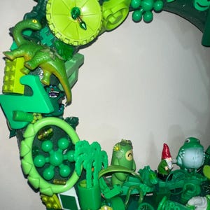 Pode incluir: Uma vibrante grinalda verde composta por vários brinquedos de plástico e elementos decorativos. A grinalda apresenta dinossauros de brinquedo, um gnomo, um sapo e outras figuras lúdicas. O design incorpora uma variedade de tons de verde, criando uma exibição caprichosa e atraente.