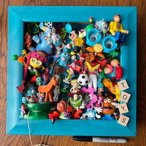 Könnte beinhalten: Ein quadratischer, blaugrüner Holzrahmen gefüllt mit einer Sammlung verschiedener Miniatur-Kunststofffiguren, darunter Zeichentrickfiguren, Tiere und ein Fußball. Vier Holzbuchstabenplättchen mit der Aufschrift TOYS liegen in der Ecke.