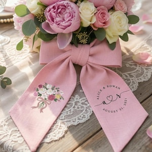 Könnte beinhalten: Ein Blumengesteck mit rosa Pfingstrosen und weißen Rosen, akzentuiert mit Grün, steht auf einem Tisch. Eine rosa Schleife mit gestickten Blumen und dem Text "Alice & Rowan August 31, 2021" ist um die Anordnung gebunden.