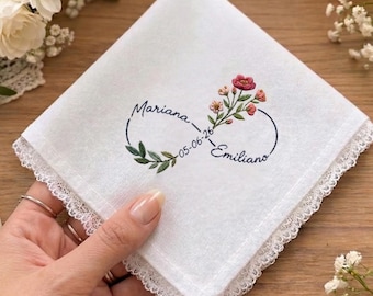 May include: A white square handkerchief with lace trim featuring an embroidered infinity symbol. The design includes the names Mariana and Emiliano, the date 05-06-26, and a small floral sprig with green leaves and pink and red flowers.