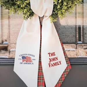 May include: A decorative wreath with a cream-colored banner. The banner has the text "UNITED STATES OF AMERICA" with an American flag and the text "THE JOHN FAMILY" in red. The banner is trimmed with red and green plaid.