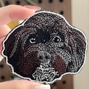 Könnte beinhalten: Gestickter Aufnäher mit einem detaillierten Hundegesicht. Der Aufnäher hat ein dunkelbraunes Fell mit hellbraunen Akzenten um Schnauze und Augen. Das Gesicht des Hundes ist von einem weißen Rand umgeben.