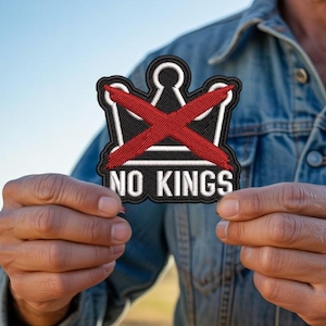 Może przedstawiać: Haftowana naszywka z czarno-białym wzorem korony, przekreślona czerwoną 'X'. Napis "NO KINGS" jest wyhaftowany na biało. Naszywka jest trzymana na tle dżinsowej kurtki i pola.