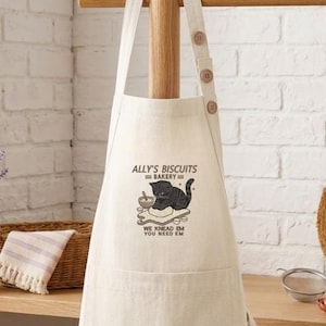 May include: A beige apron featuring a graphic of a black cat kneading dough with the text Ally's Biscuits Bakery We Knead 'Em You Need 'Em. The apron has a neck strap with three visible buttons and a large front pocket for utility and storage.