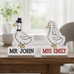 以下が含まれることがあります： 刺繍された結婚式のガチョウの置物。片方はシルクハットと蝶ネクタイ、もう片方はベールをかぶっています。それぞれにネームプレートがあり、「MR JOHN」と「MRS EMILY」と書かれています。置物は木製のテーブルの上にあります。