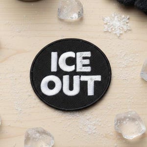 Op de afbeelding: Een zwarte, ronde patch met de woorden "ICE OUT" in witte blokletters. De patch is omgeven door ijsblokjes, sneeuwvlokken, een metalen mok en een zwarte handschoen op een licht houten oppervlak.