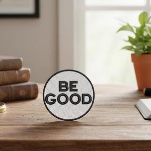 Peut inclure: Un écusson rond brodé avec les mots "BE GOOD" en lettres noires sur fond blanc. L'écusson a une bordure noire. L'écusson est posé sur un bureau en bois avec des livres, une boussole et une plante en pot en arrière-plan.