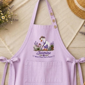 Puede incluir: Un delantal de color morado claro con un gráfico central de un pato con sombrero y peto, sosteniendo una herramienta de jardín. El texto dice Jasmine y Bloom Where You're Planted. El delantal incluye un bolsillo frontal, tirantes con botones y lazos.