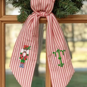 Puede incluir: Un lazo de tela a rayas rojas y blancas con una corona navideña. El lazo tiene un cascanueces bordado en un lado y una letra "T" bordada en verde en el otro. El lazo está atado en el centro.