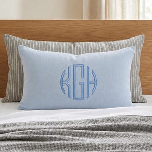 May include: A rectangular light blue throw pillow featuring a centered circular monogram with the letters KGH embroidered in a matching blue thread. The pillow is positioned in front of a larger gray and white striped pillow on a bed.