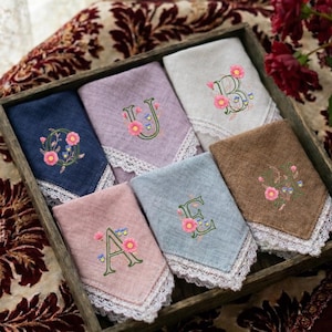 Può includere: Un set di sei tovaglioli di stoffa quadrati in blu navy, lavanda, crema, rosa, azzurro e marrone, ognuno con una lettera ricamata decorata con motivi floreali rosa e blu e rifinito con un bordo in pizzo bianco.
