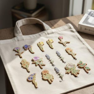 Puede incluir: Un bolso tote beige con una variedad de parches de flores bordadas en varios colores. Los parches incluyen iris, rosas y lavanda. La bolsa está sobre una superficie de madera, con un libro visible en el fondo.