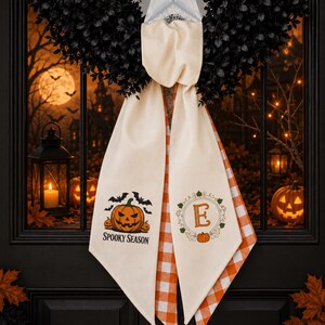 Puede incluir: Un colgador de puerta de tela color crema con dos extremos colgantes. El extremo izquierdo presenta una calabaza, murci&eacute;lagos y el texto "SPOOKY SEASON". El extremo derecho muestra una letra "E" dentro de una corona floral con una calabaza.