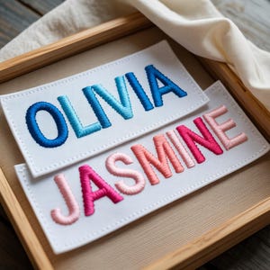 Puede incluir: Dos parches rectangulares blancos con nombres bordados. El parche superior dice "OLIVIA" en hilo degradado azul. El parche inferior dice "JASMINE" en hilo degradado rosa y coral. Los parches se muestran en una bandeja de madera.