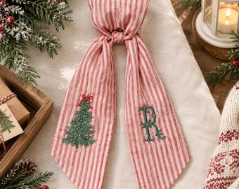May include: A red and white striped fabric hair accessory featuring an embroidered green Christmas tree with a red bow on one side and a decorative green embroidered letter R on the other side, tied in a knot at the top with two long tails.