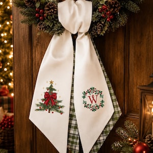 May include: A white fabric hanging decoration featuring a Christmas tree with a red bow on the left panel and a green wreath with a red letter W in the centre on the right panel. The back side of the fabric displays a green and white plaid pattern.