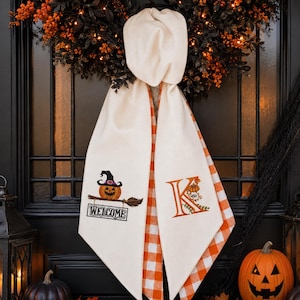 May include: A cream-colored fabric door hanger featuring a top knot and two hanging panels. The left panel displays an embroidered pumpkin wearing a witch hat on a broomstick above the text "WELCOME". The right panel features a large orange letter K.