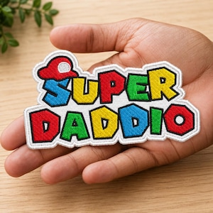 May include: Embroidered patch with the words "SUPER DADDIO" in a colorful, cartoon-style font. The patch features a red, blue, green, and yellow color scheme with a white border. The patch is held in a person's hand.