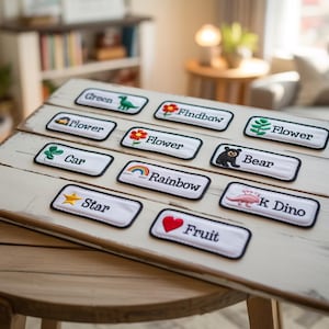 May include: A wooden sign displaying embroidered patches with black borders. Each patch has a word and image, such as a dinosaur, flower, bear, and rainbow. The sign rests on a round wooden table, with a neutral background.