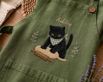 May include: An olive green apron featuring an embroidered black cat wearing a checkered neckerchief, kneading dough with a rolling pin. The name Ashley is embroidered above the cat, and the apron includes three brown buttons on the shoulder strap.
