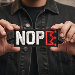 May include: A rectangular patch with the word "NOPE" in white, textured lettering. The "E" is replaced with a red crown design. The patch is held in front of a person wearing a black denim jacket.