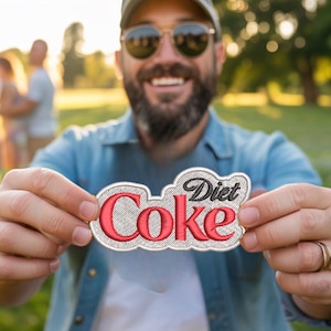 Puede incluir: Parche bordado con las palabras "Diet Coke" en rojo y negro. El parche tiene una apariencia texturizada y cosida. El parche se sostiene frente a una persona. El fondo es una escena borrosa al aire libre.
