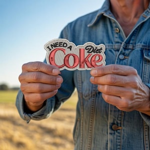 May include: Embroidered patch with the words "I Need a Diet Coke" in red and black lettering. The patch is held in front of a denim jacket. The patch has a white border.