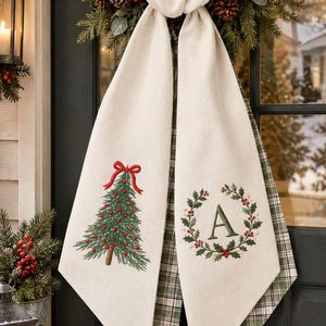 May include: A cream-colored fabric door hanger featuring two long, pointed panels. One panel displays an embroidered green Christmas tree with a red bow, while the other features a green holly wreath surrounding the letter A.