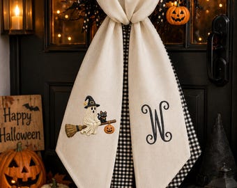 May include: A cream-colored fabric door hanger featuring a black and white gingham lining. One side displays an embroidered ghost wearing a witch hat, riding a broom with a black cat and pumpkin. The other side features a large, black embroidered letter W.