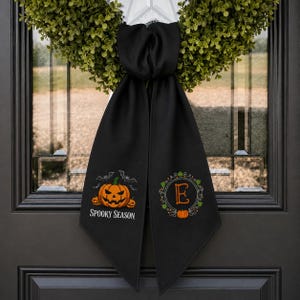 Puede incluir: Un colgador de puerta de tela negra con una corona verde en la parte superior. El lado izquierdo muestra una calabaza naranja con murci&eacute;lagos y el texto SPOOKY SEASON. El lado derecho presenta un marco circular decorativo con la letra E.