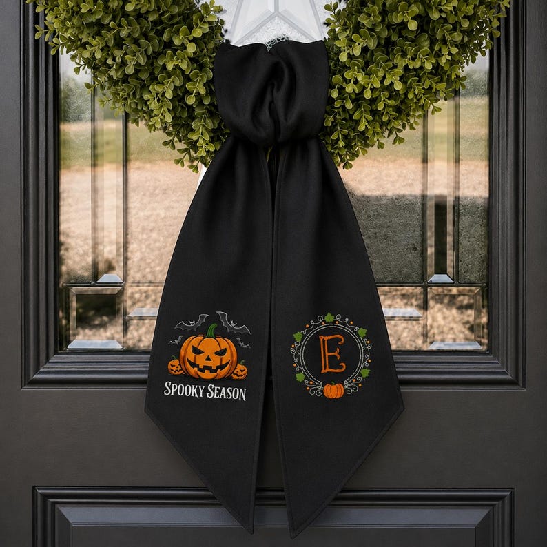 Puede incluir: Un colgador de puerta de tela negra con una corona verde en la parte superior. El lado izquierdo muestra una calabaza naranja con murci&eacute;lagos y el texto SPOOKY SEASON. El lado derecho presenta un marco circular decorativo con la letra E.