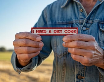 Puede incluir: Un parche rectangular con el texto "I NEED A DIET COKE" en letras rojas. El parche lo sostiene una persona que lleva una chaqueta vaquera. El fondo muestra un campo y un cielo azul.