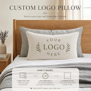 May include: A rectangular off-white throw pillow featuring centered dark green embroidery that reads YOUR LOGO HERE, flanked by two leafy sprigs. The pillow rests on a bed with a gray striped pillowcase and a gray textured blanket.