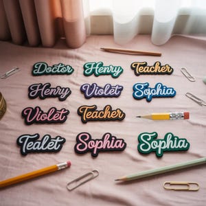 May include: A collection of name tags with various names like "Doctor", "Henry", "Teacher", and "Sophia" in a cursive font. The tags have a black border with colorful lettering. Pencils and paper clips are scattered around the tags on a pink surface.