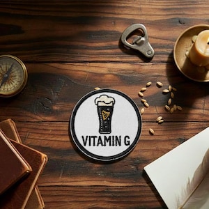May include: A circular patch with a black and white design of a pint of beer, with the words "VITAMIN G". A bottle opener, compass, candle, and grains are also visible. The patch has a black border.