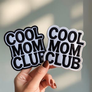 Pode incluir: Dois patches bordados com o texto "COOL MOM CLUB". Um é preto com letras brancas e o outro é branco com letras pretas. Os patches têm uma borda ondulada.